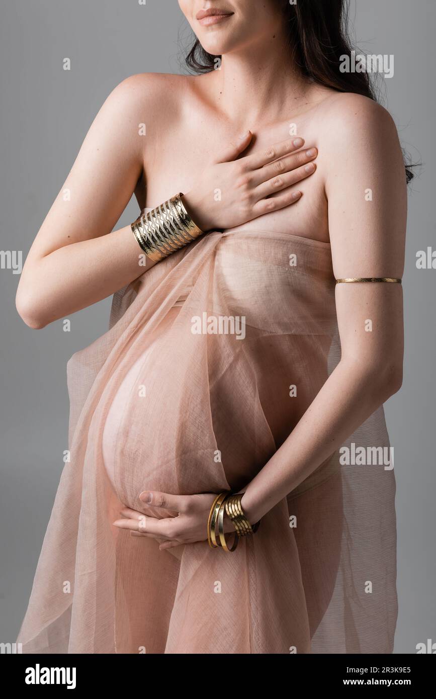 cropped view of stylish and elegant pregnant woman posing in golden accessories and transparent chiffon draping isolated on grey background, maternity Stock Photo