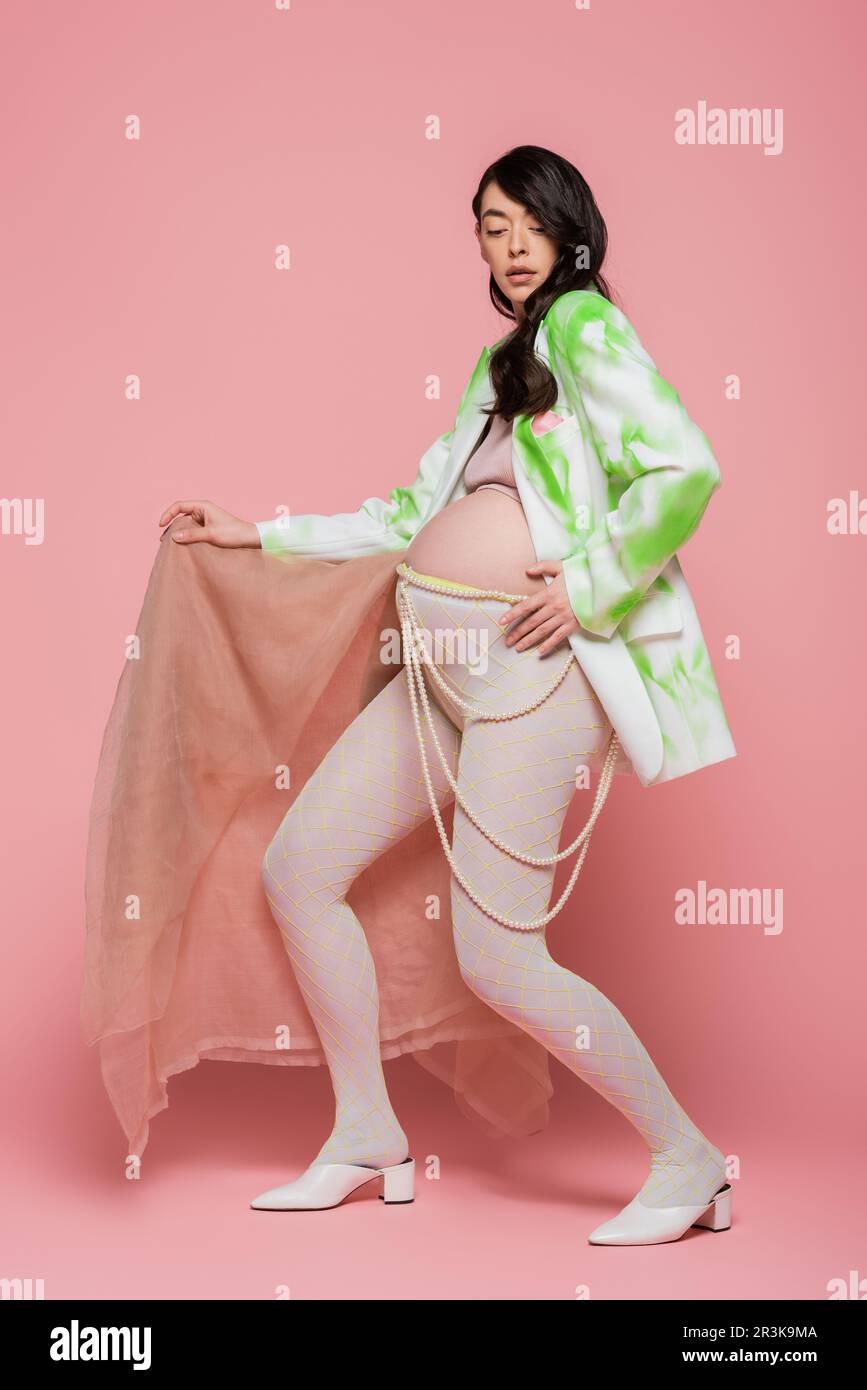 full length of brunette pregnant woman in trendy blazer, crop top, beads belt and leggings posing with beige chiffon cloth on pink background, materni Stock Photo