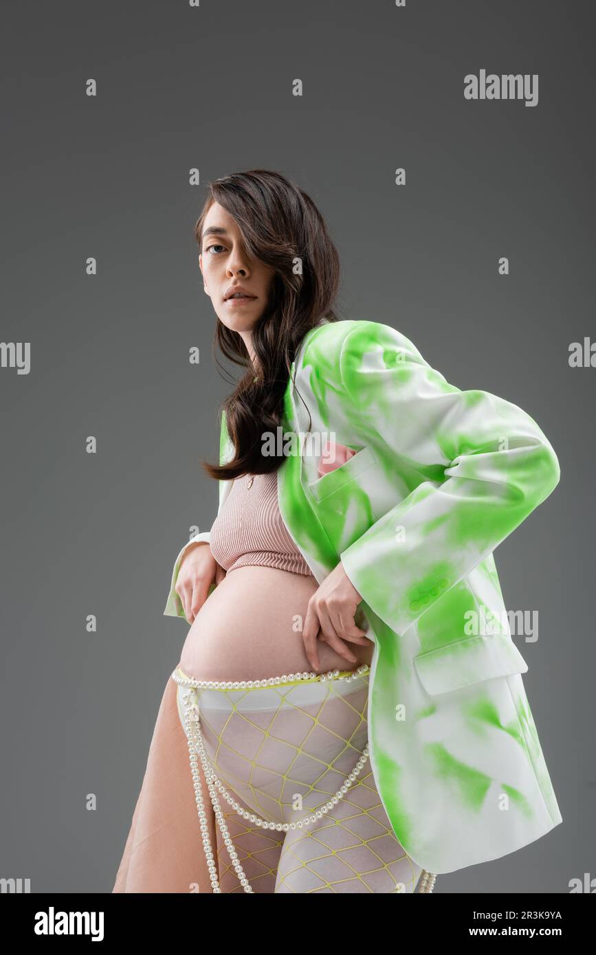 brunette pregnant woman in stylish jacket, crop top, beads belt and leggings with chiffon cloth looking at camera isolated on grey background, materni Stock Photo