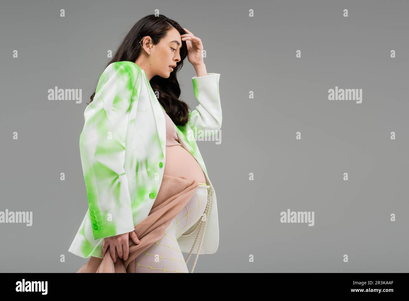 side view of pretty pregnant woman in green and white jacket and leggings with beads belt and beige chiffon cloth isolated on grey background, materni Stock Photo