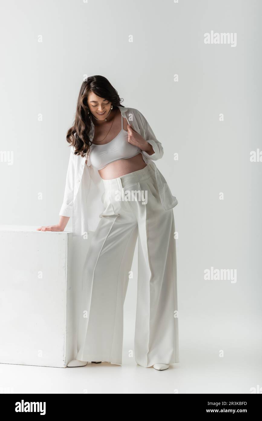 full length of pleased pregnant woman with wavy brunette hair, wearing white pants, crop top and shirt, standing near cube on grey background, materni Stock Photo