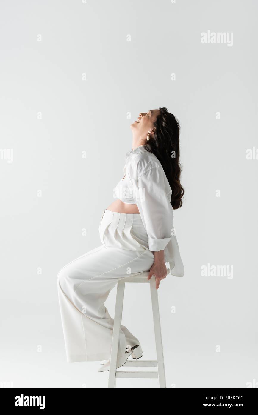 side view of overjoyed pregnant woman with wavy brunette hair sitting on stool in white pants, shirt and crop top isolated on grey background, materni Stock Photo