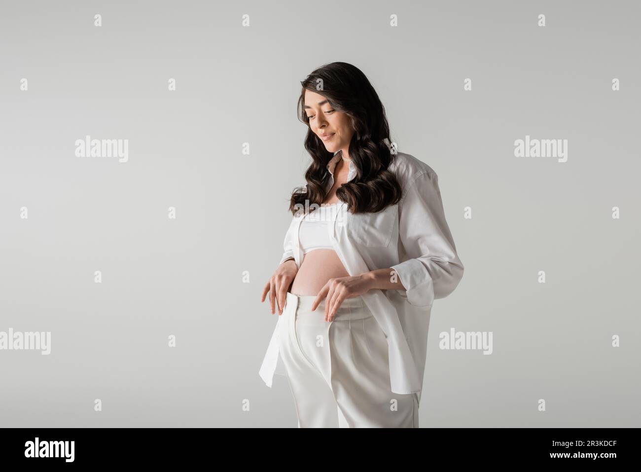 charming and happy pregnant woman with wavy brunette hair posing in white shirt, crop top and pants while smiling isolated on grey background, materni Stock Photo