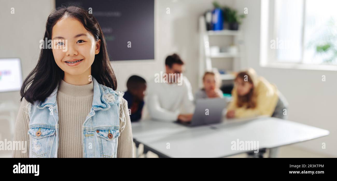 A confident student looks at the camera while the teacher engages other students in the background. Girl attending a digital literacy class at school. Stock Photo