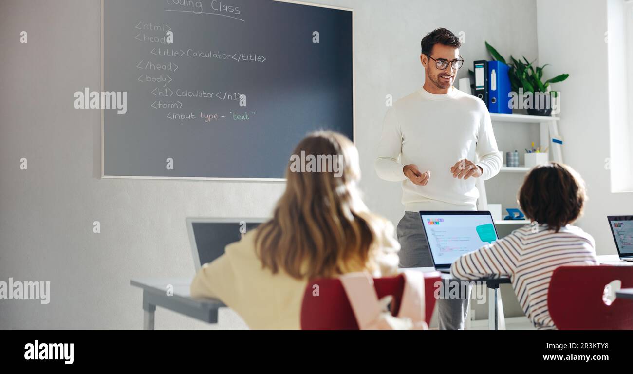 Male teacher guiding children through learning the process of coding. Standing in front of the class, he explains the fundamentals of programming and Stock Photo