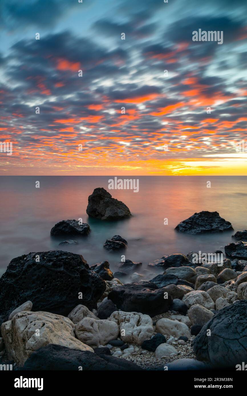 A colorful sunrise at Cala Gonone with black and white rocks and boulders in the foreground Stock Photo