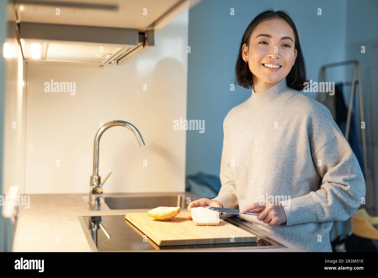 Cute asian woman making herself toast, cut loaf of bread, preparing sandwitch on kitchen Stock Photo
