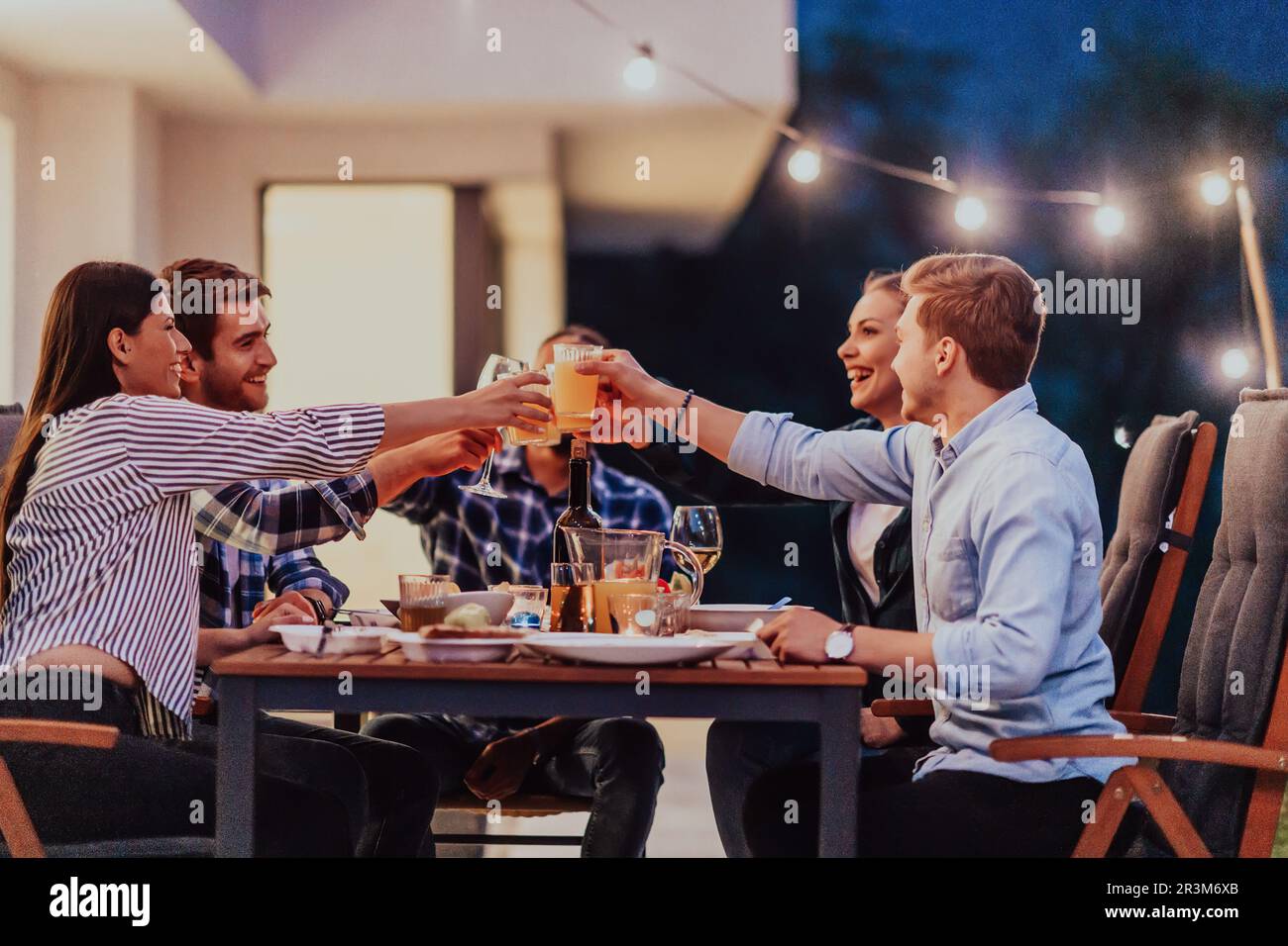 A group of young diverse people having dinner on the terrace of a modern house in the evening. Fun for friends and family. Celeb Stock Photo