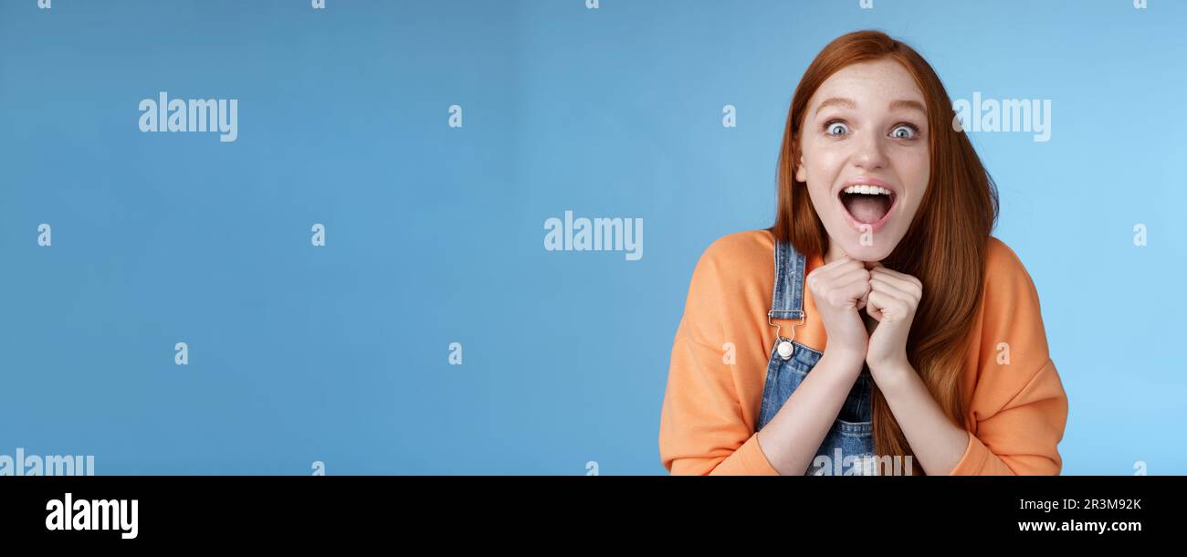 Cute redhead european girl blue eyes freckles reacting amused shocking rumor lift eyebrows drop jaw surprised smiling excited pi Stock Photo
