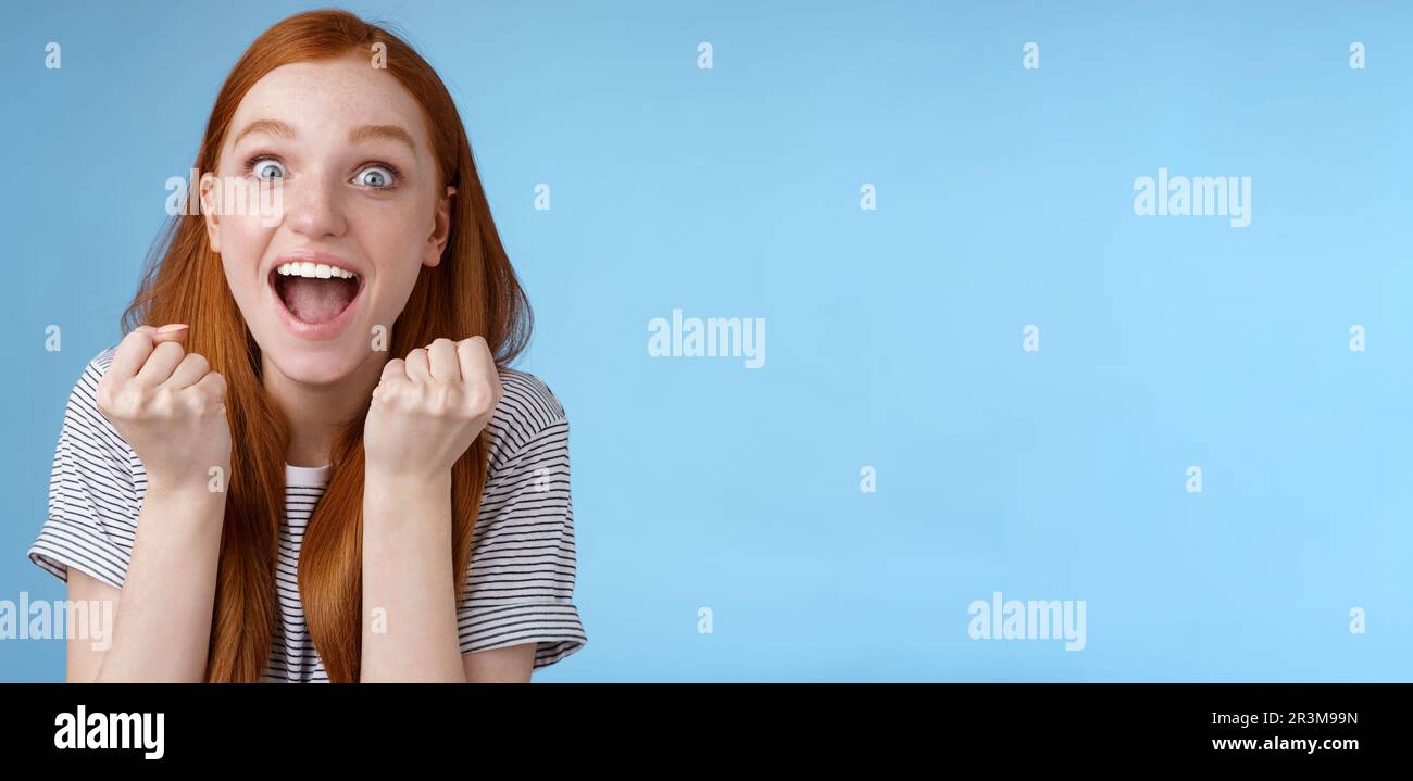 Yes awesome news great. Attractive surprised redhead girl cheering happy clench fists yelling yeah accomplish goal wide eyes exc Stock Photo