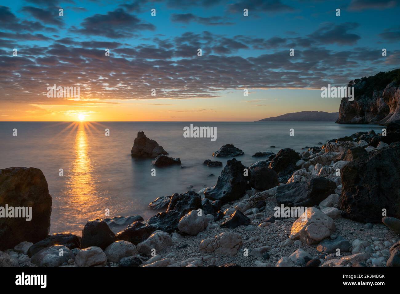 A colorful sunrise at Cala Gonone with black and white rocks and boulders and a sunburst Stock Photo