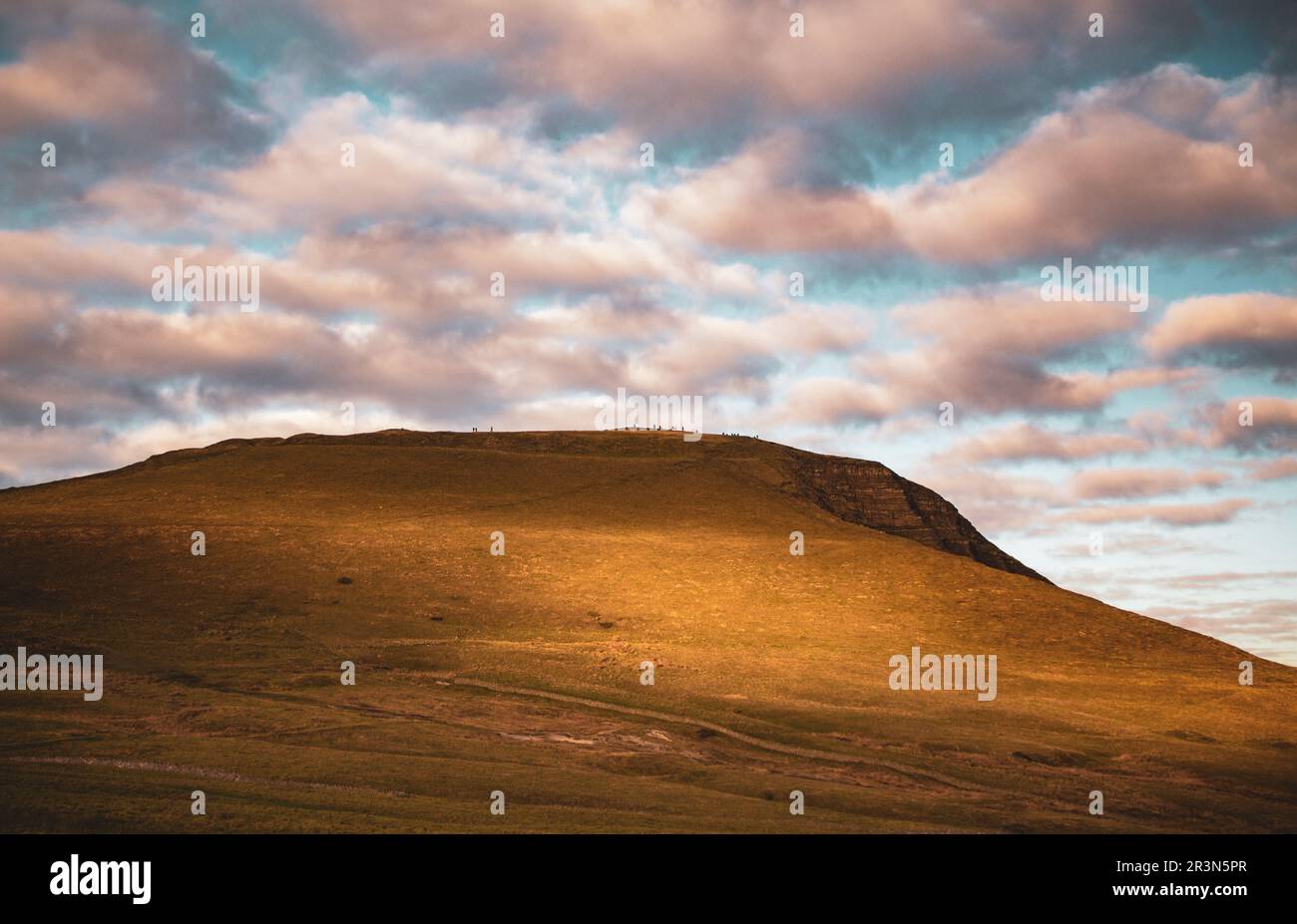 Wide shot of Mam Tor, a landmark hill in the English Peak District washed in a golden sunset with a dramatic cloudy sky and hikers far into the distan Stock Photo