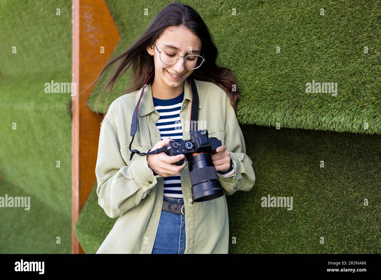 Young female photographer in casual clothes smiling and looking at photo camera while standing near green wall Stock Photo