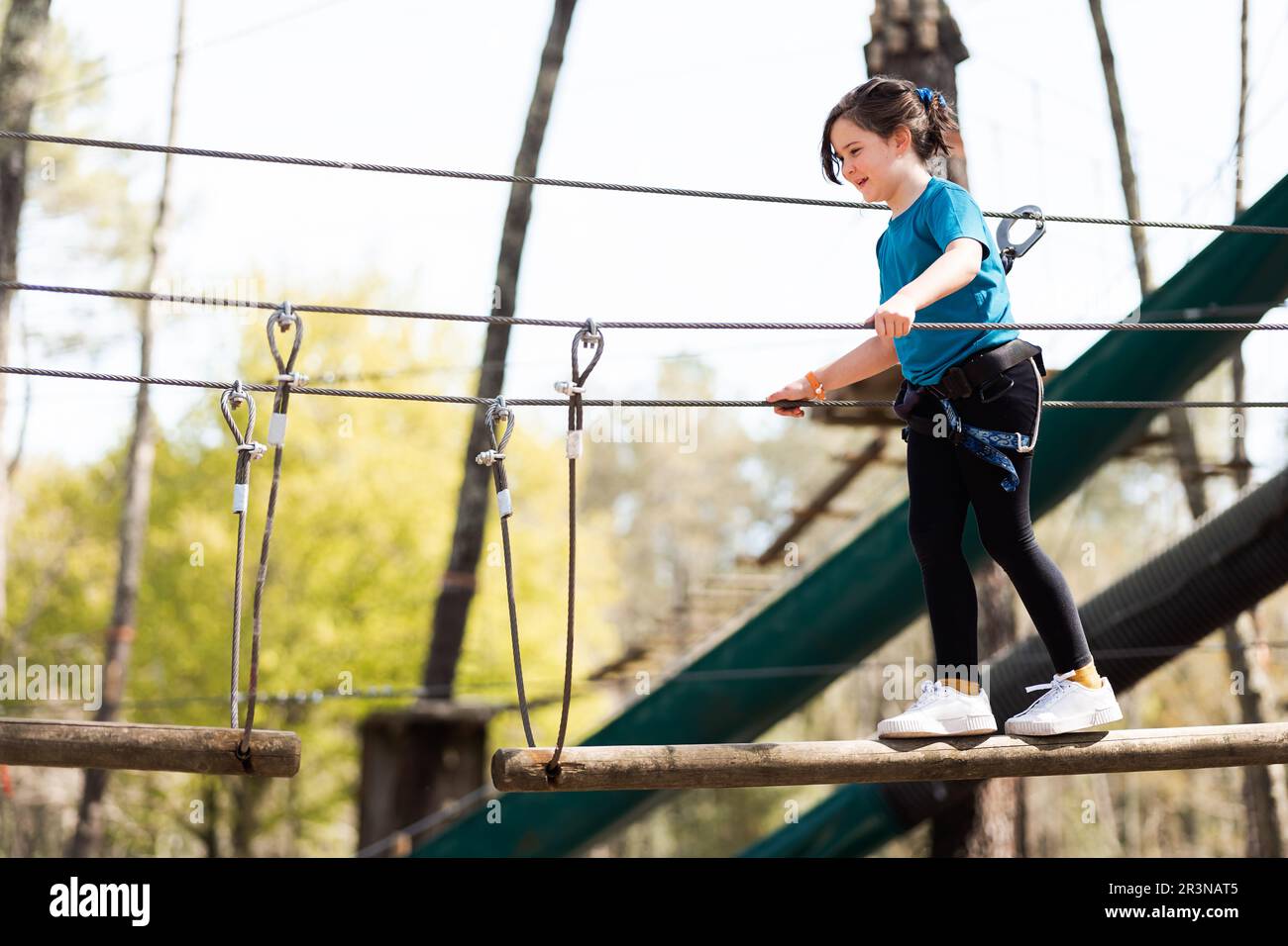 From below full body of a happy and focused preteen girl in casual clothes and safety gear standing balancing while walking on a wooden bridge in the Stock Photo