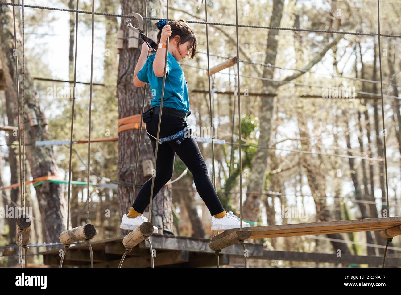 Side view of and focused preteen girl in casual clothes and safety gear standing balancing while walking on a wooden bridge in the rope park Stock Photo