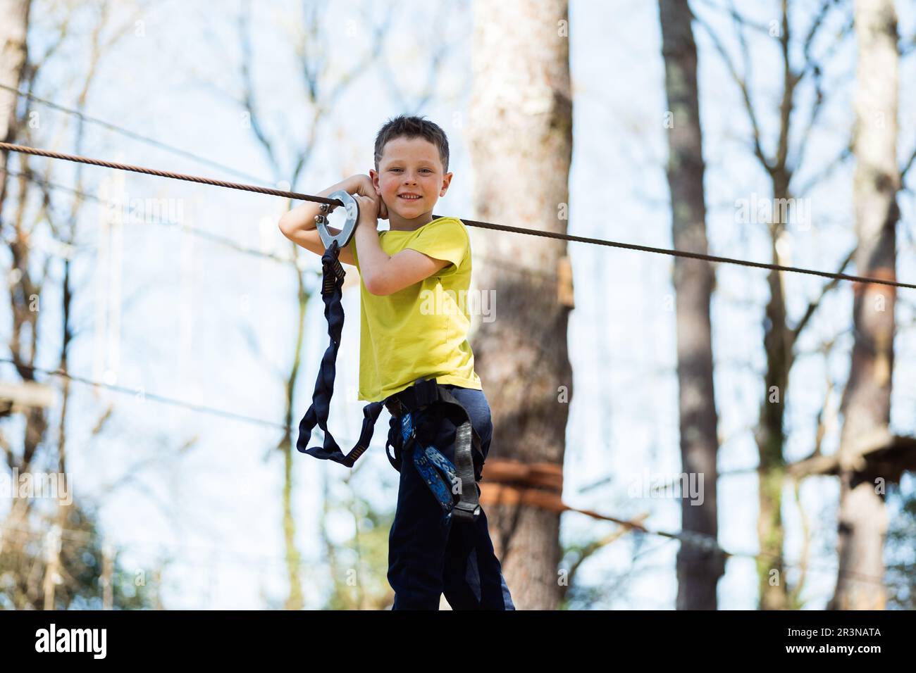 From below cheerful preteen boy in casual clothes and safety gear standing holding on to the rope of a wooden bridge and looking at the camera as he p Stock Photo