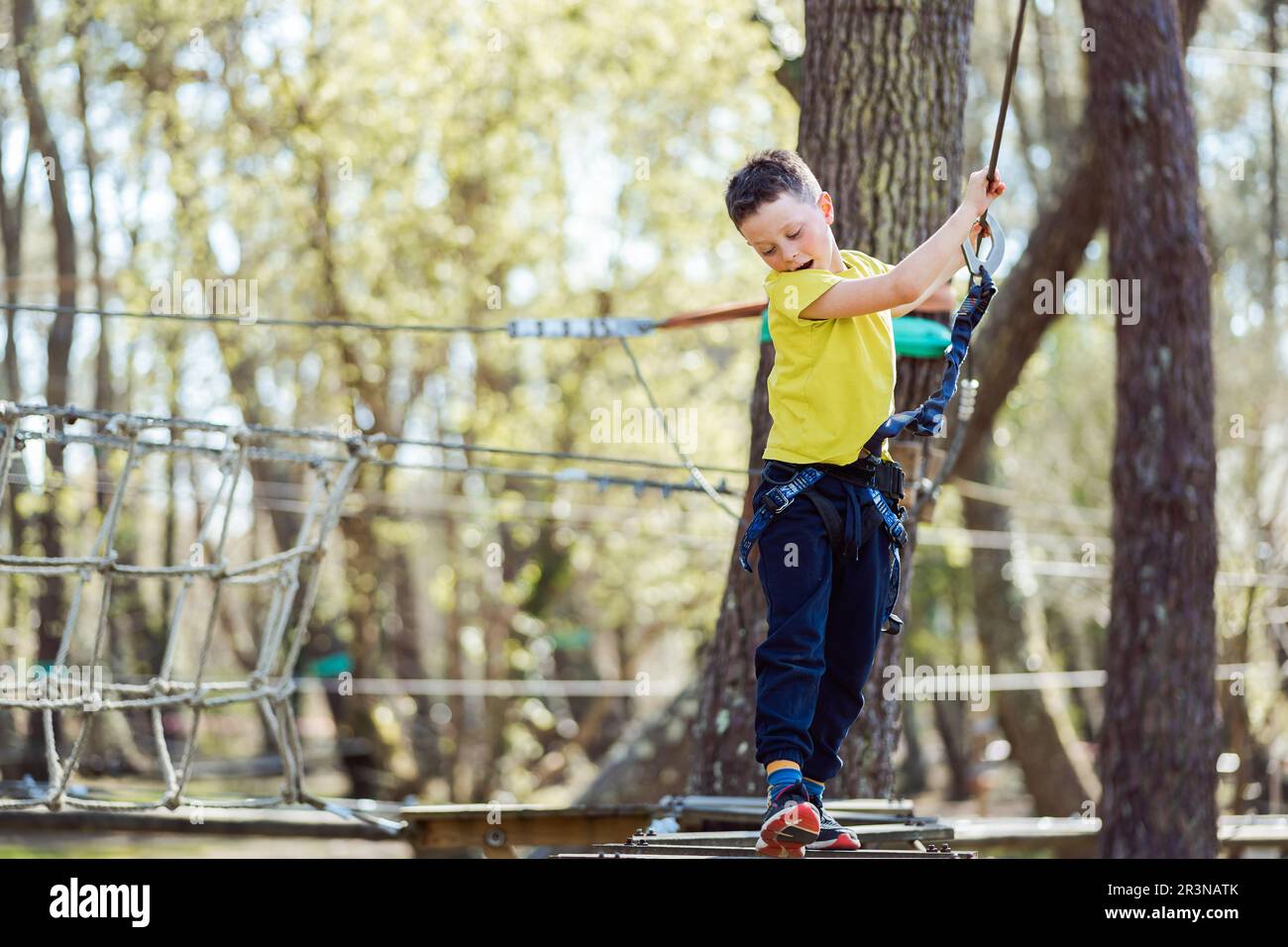 Full body of an active preteen boy in casual clothes and safety gear standing on a wooden rope bridge and looking down as he passes the time in the pa Stock Photo