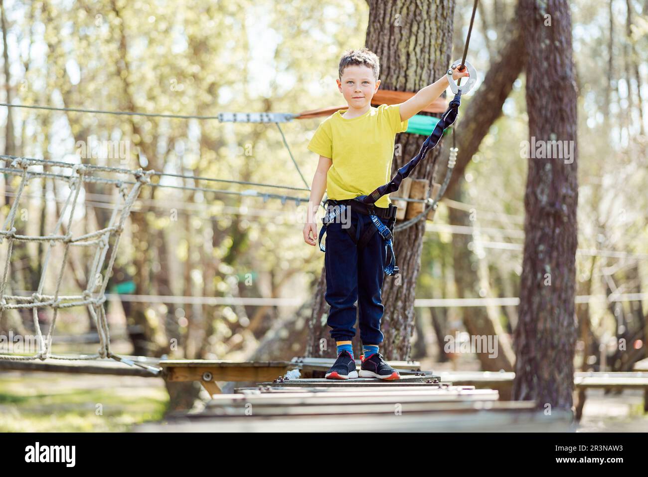 Full body of active preteen boy in casual clothes with safety equipment standing on wooden rope bridge and looking at camera while spending time in pa Stock Photo