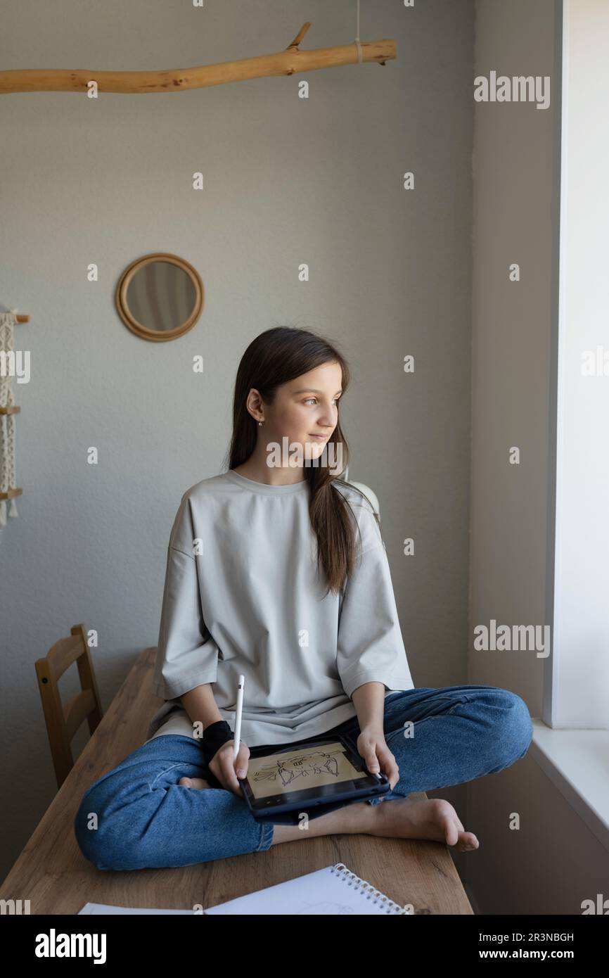 Positive teen girl sitting with legs crossed on table and drawing on a graphic tablet while looking away in her own room Stock Photo