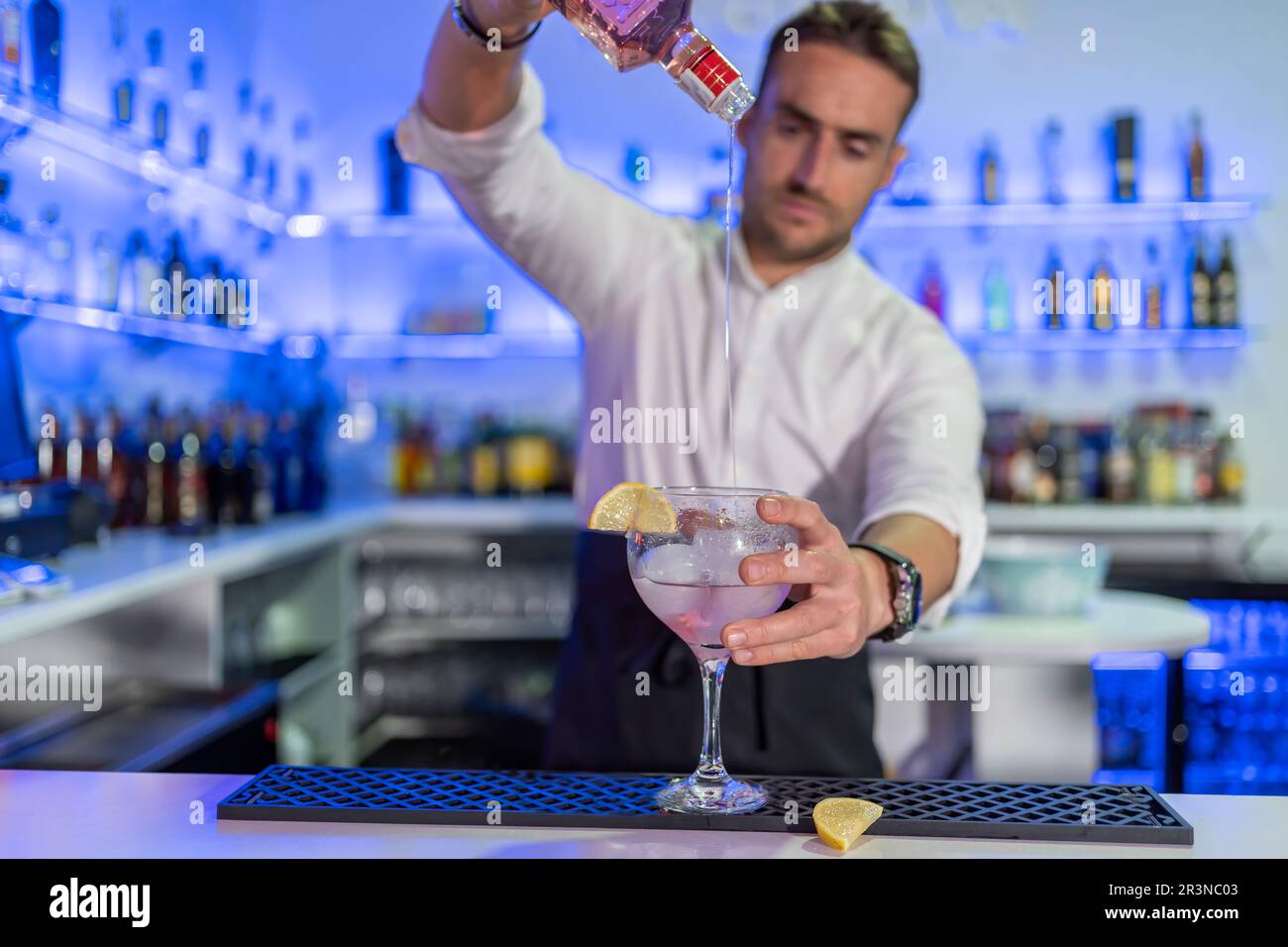 Focused young male barkeeper pouring alcohol drink from bottle into glass of cocktail with orange slice on counter while looking down against blurred Stock Photo
