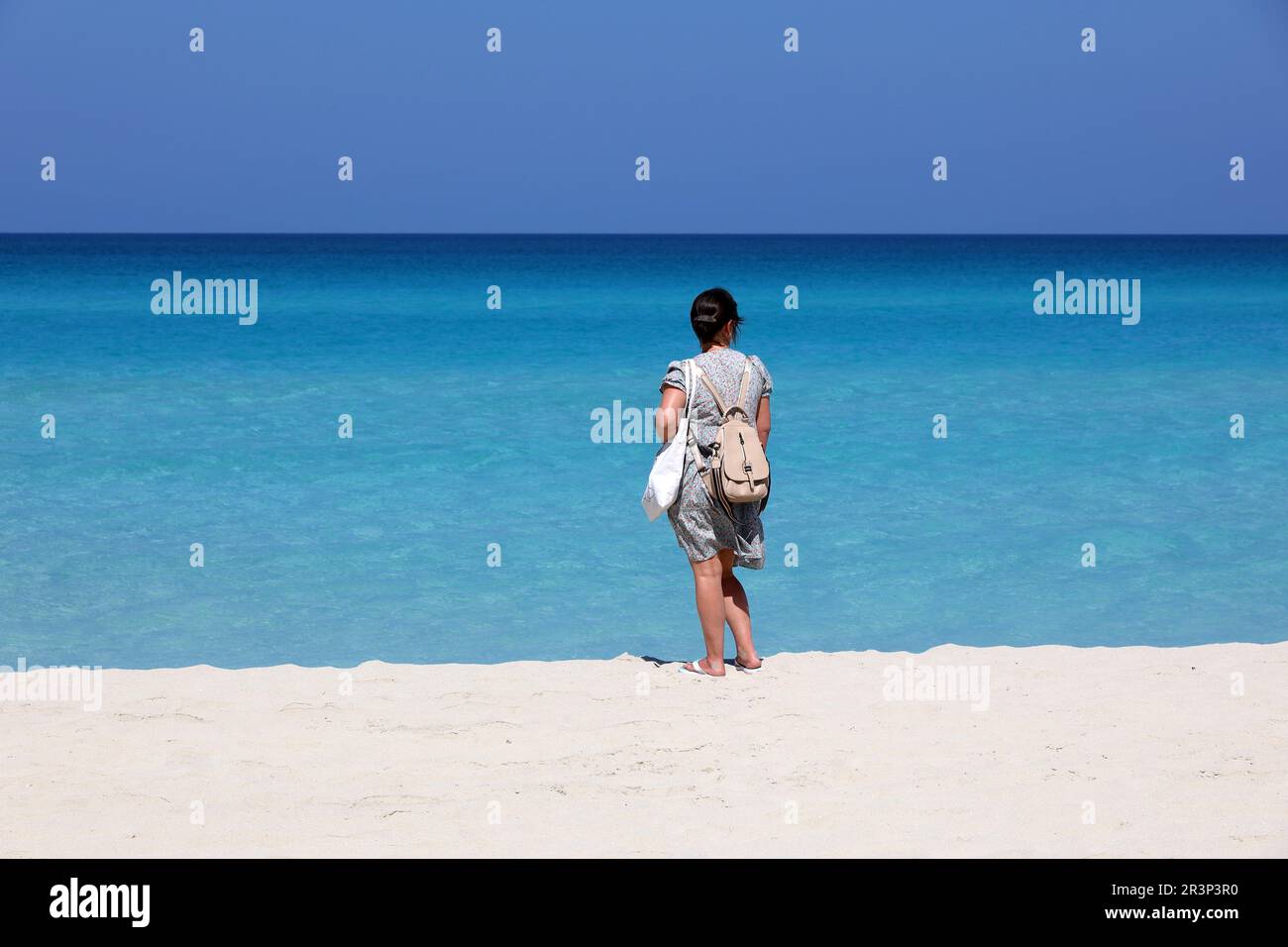 Woman in summer dress standing with backpack purse on white sand against the azure waves. Travel and beach vacation on ocean coast Stock Photo