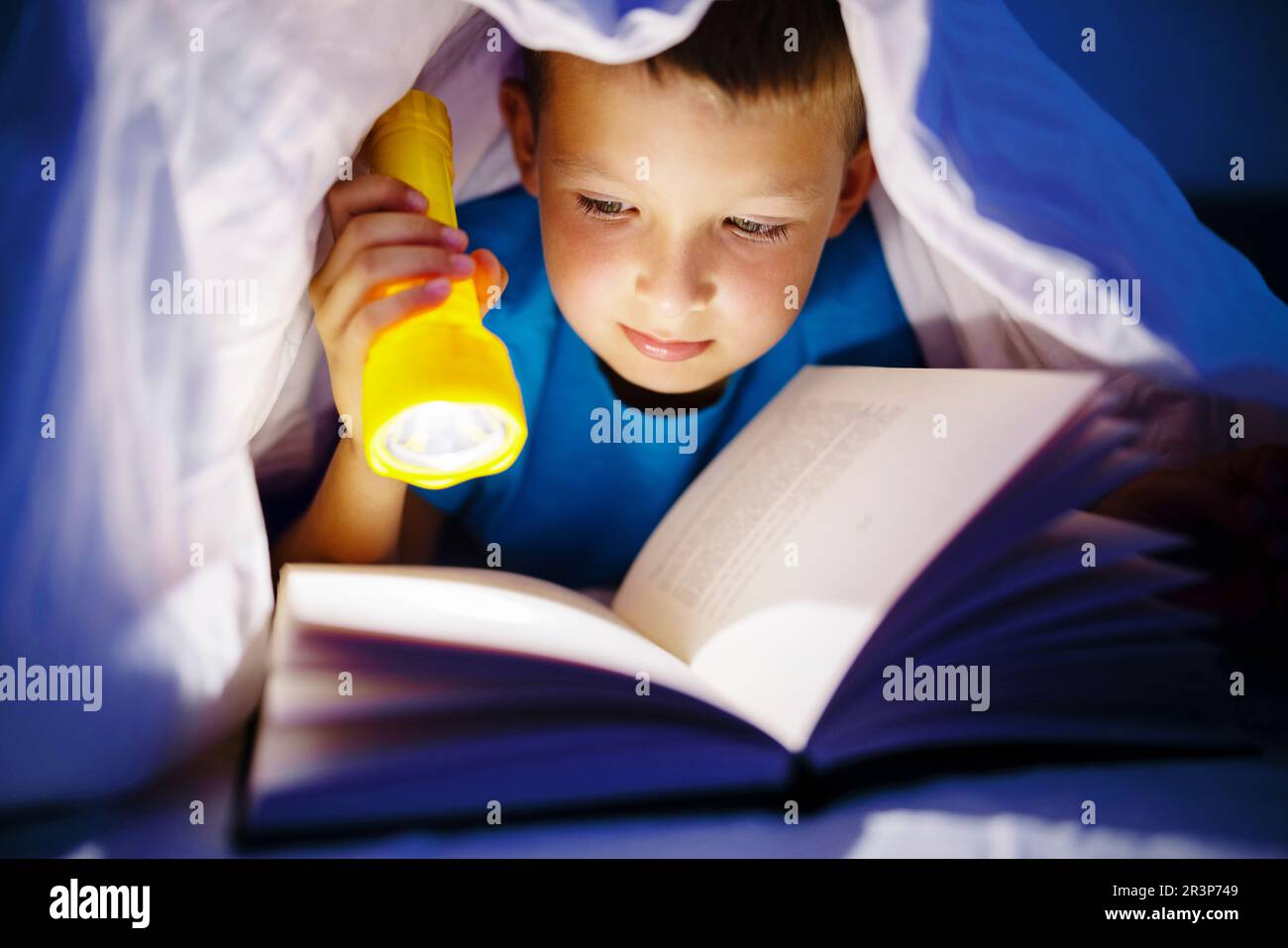 A young boy reading a book under the covers with a flashlight at dark