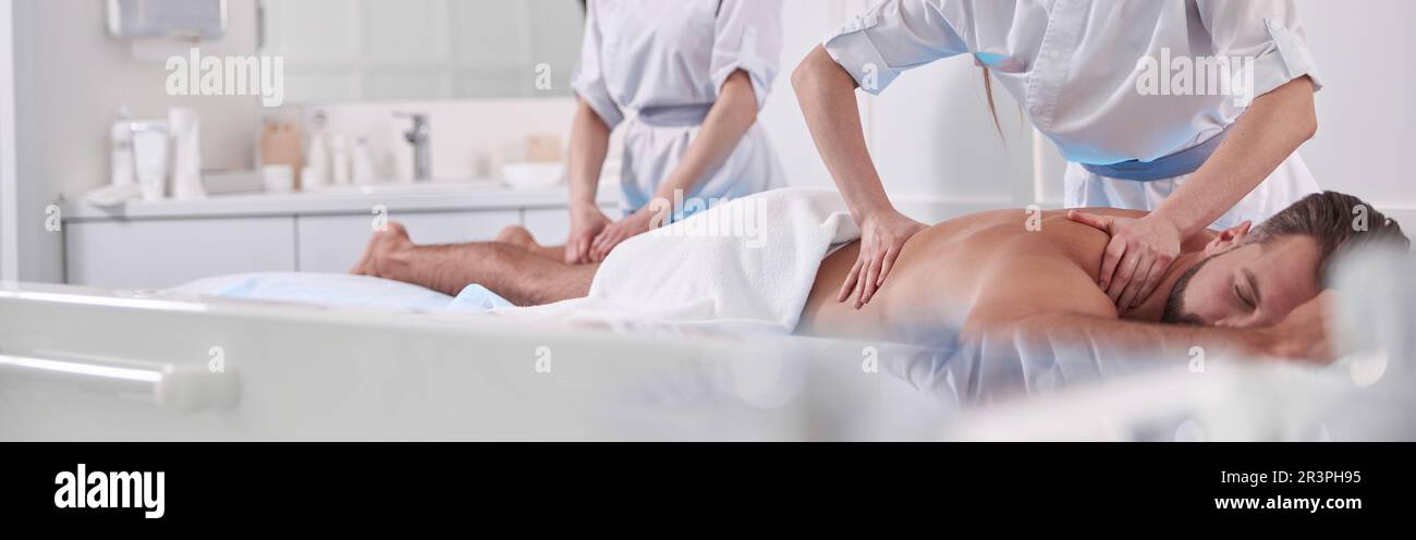 Woman does massage of back to man while colleague works with his leg on couch in hospital Stock Photo