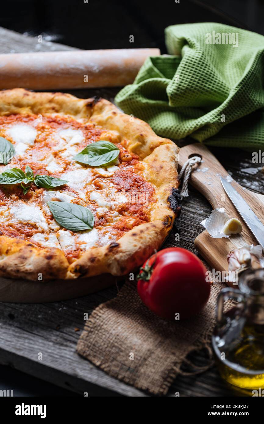 Italian Pizza Napoletana in Messy Kitchen with Cooking Ingredients such as tomato, olive oil, garlic, basil, rolling pin, flour, knife, cutting board Stock Photo