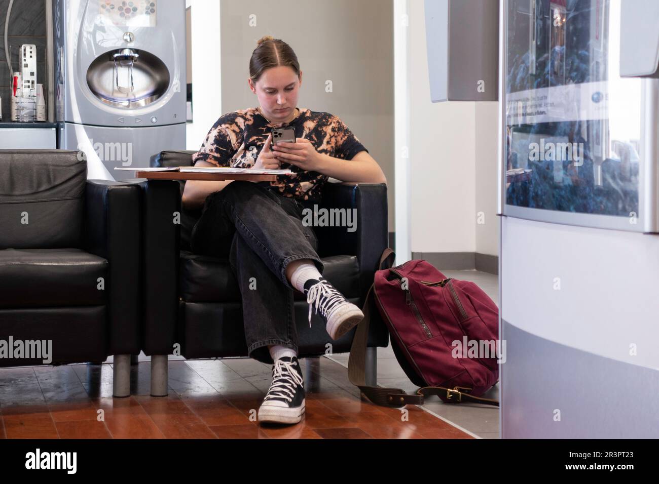 A young Caucasian woman with a red backpack in an automobile service waiting room looking at her mobile. Kansas, USA. Stock Photo