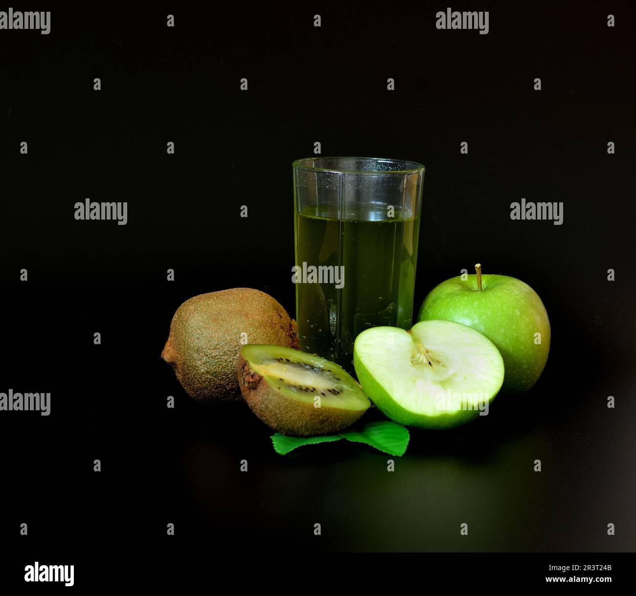 Fruit juice in a tall glass and pieces of ripe green apple and kiwi with leaves on a black background. Close-up. Stock Photo