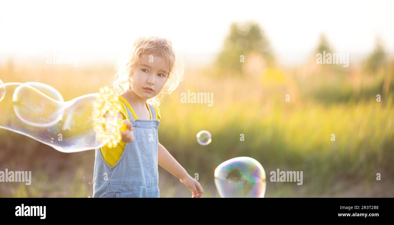 A girl in a denim jumpsuit blows soap bubbles in the summer in a field at sunset. International Children's Day, happy child, out Stock Photo