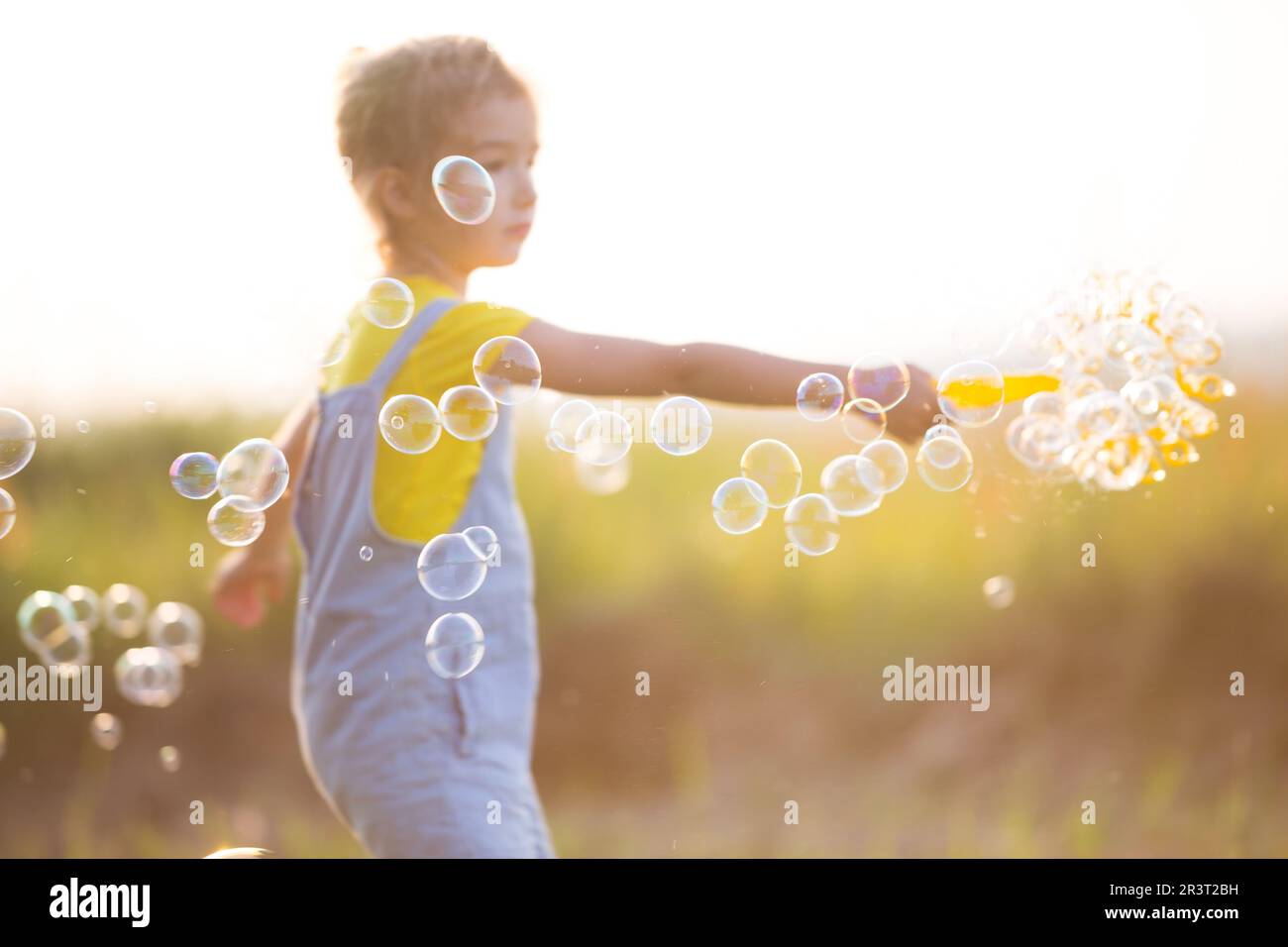 A girl in a denim jumpsuit blows soap bubbles in the summer in a field at sunset. International Children's Day, happy child, out Stock Photo