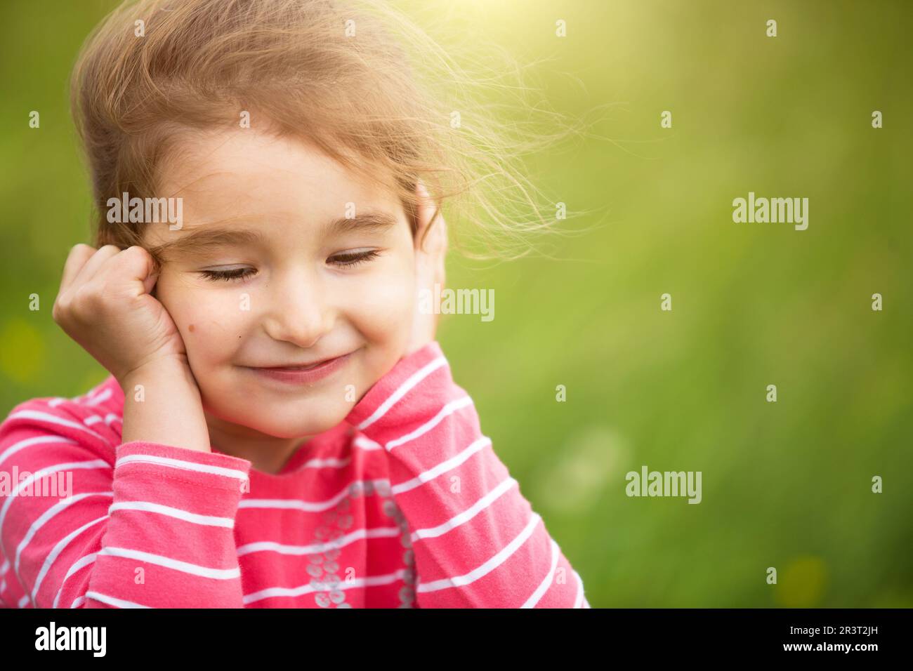 Little girl in a coral striped T-shirt on a green background in a field holds her face in her hands and smiles slyly. Children's Stock Photo