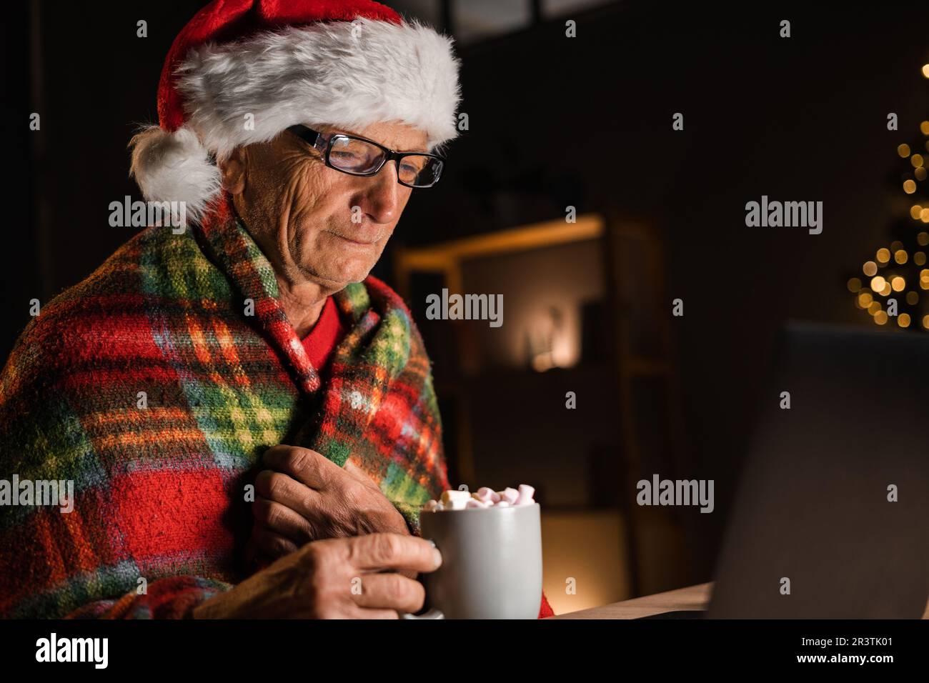Side view of old man drinking cocoa with marshmallows on christmas eve, Elderly man in santa hat alone watching movie on internet at home Stock Photo