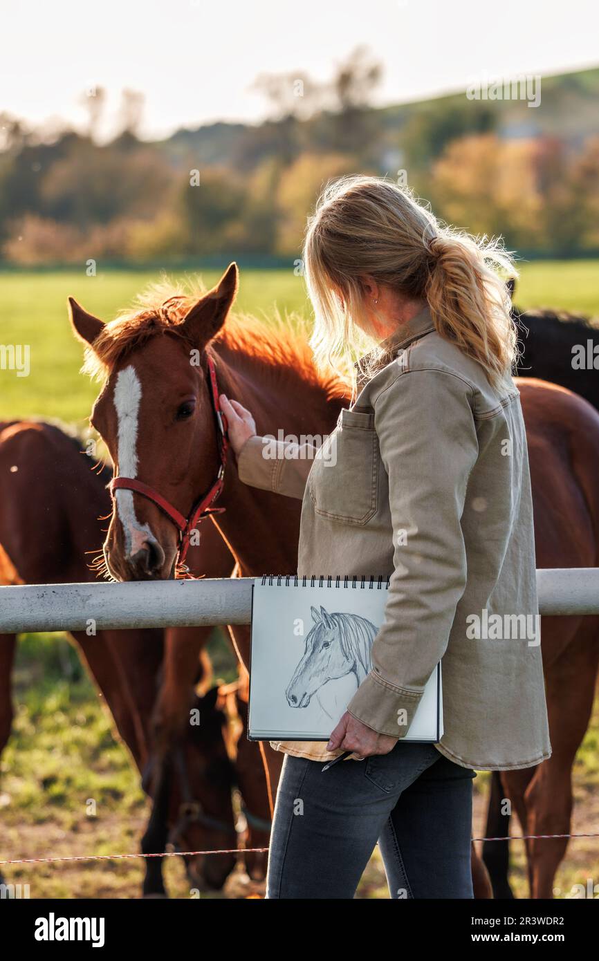 Female artist holding sketchbook with pencil drawing. Woman relax with horses on pasture Stock Photo