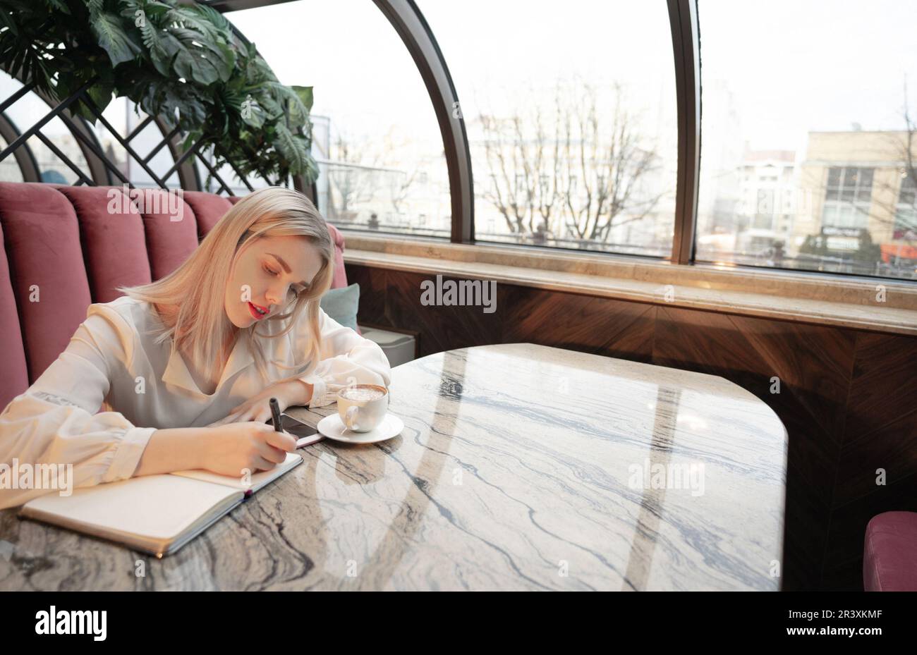 Dreamy young woman writing down future plans and goals in own diary resting in cozy cafeteria Stock Photo