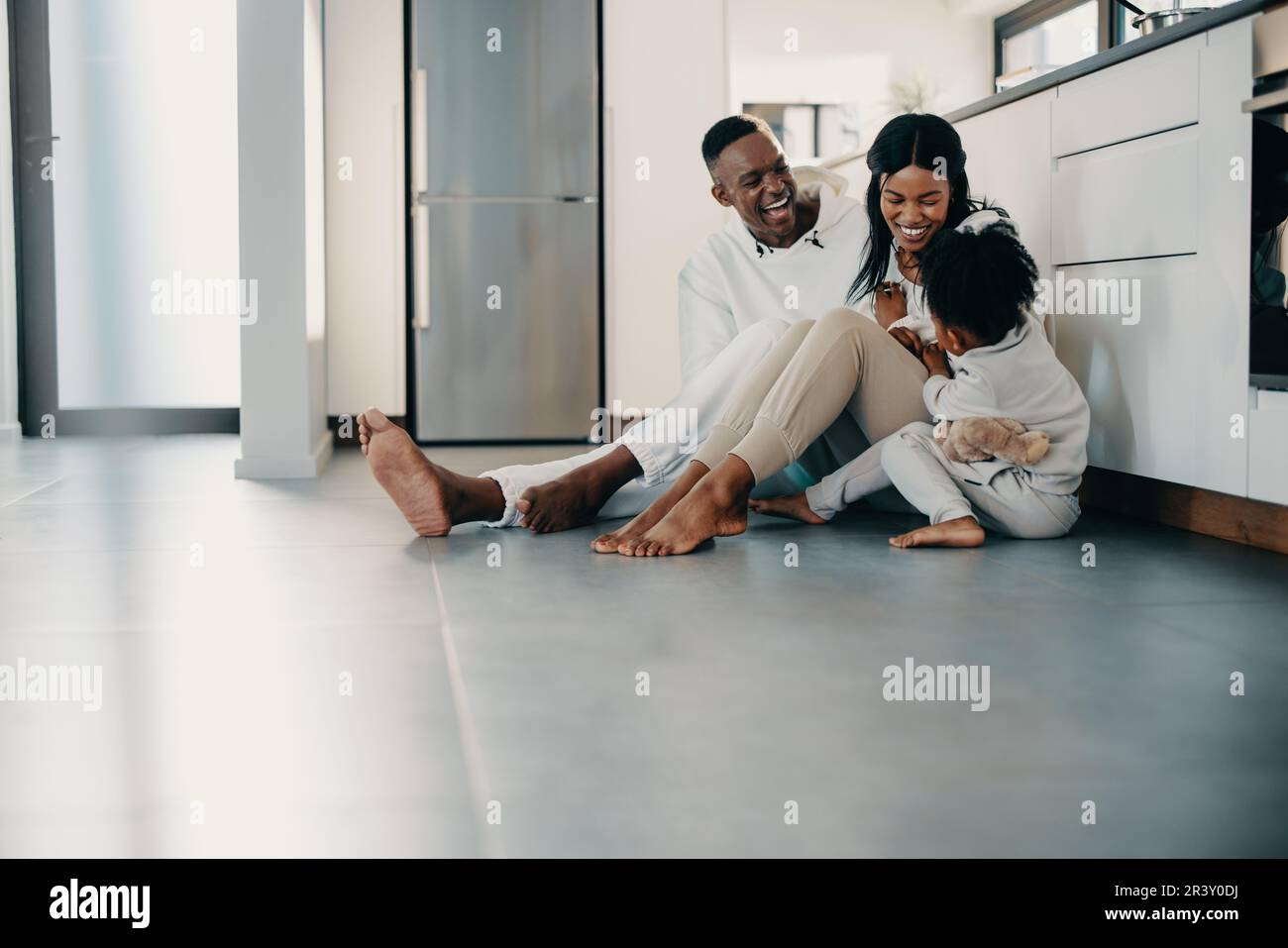 Family of three playing together and enjoying a fun moment. Parents having fun with their daughter while sitting on the floor in the kitchen. Black co Stock Photo