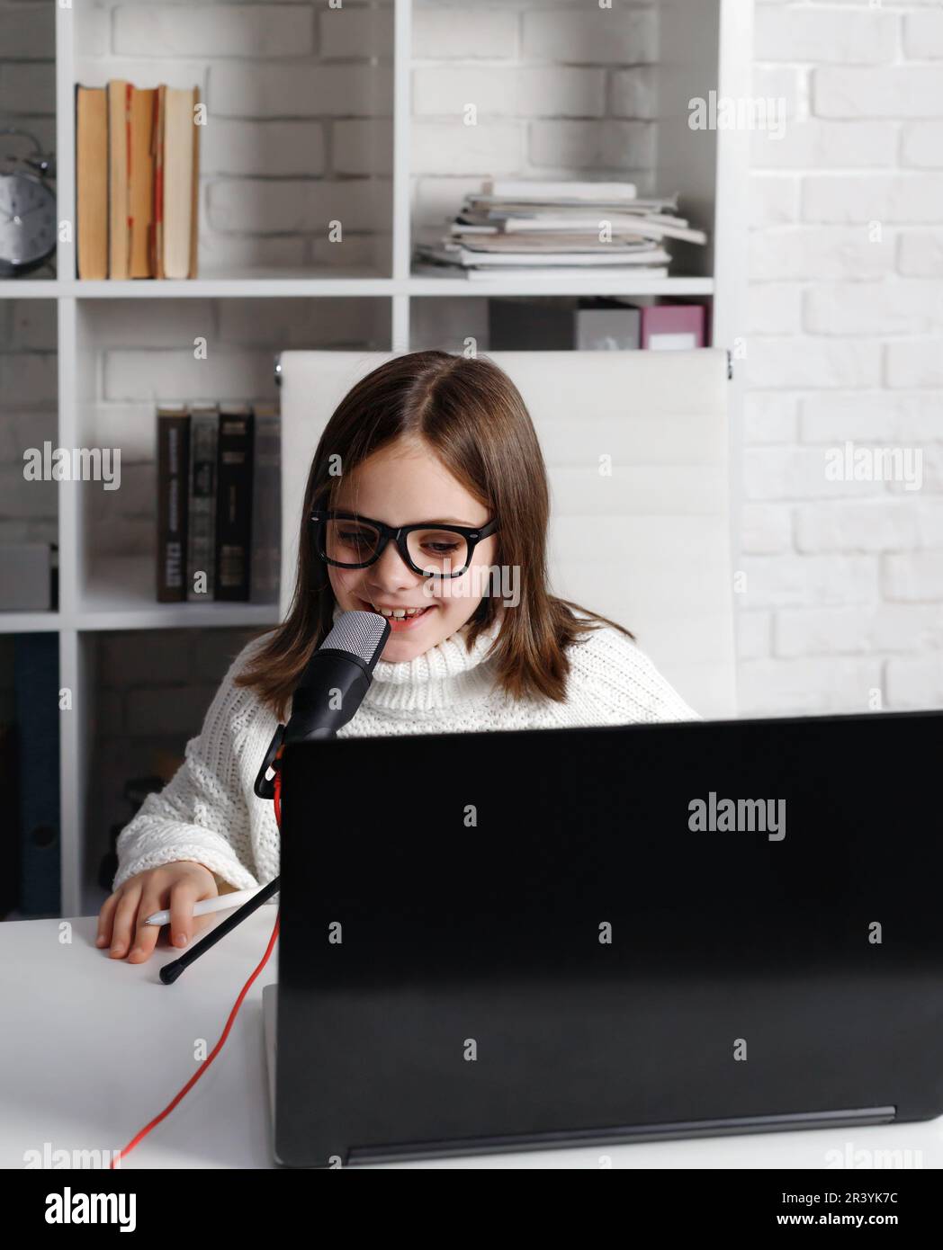 Little girl in eyeglasses sitting at white table, kid - blogger smiling and speaking into microphone, child using laptop and looking at screen while r Stock Photo