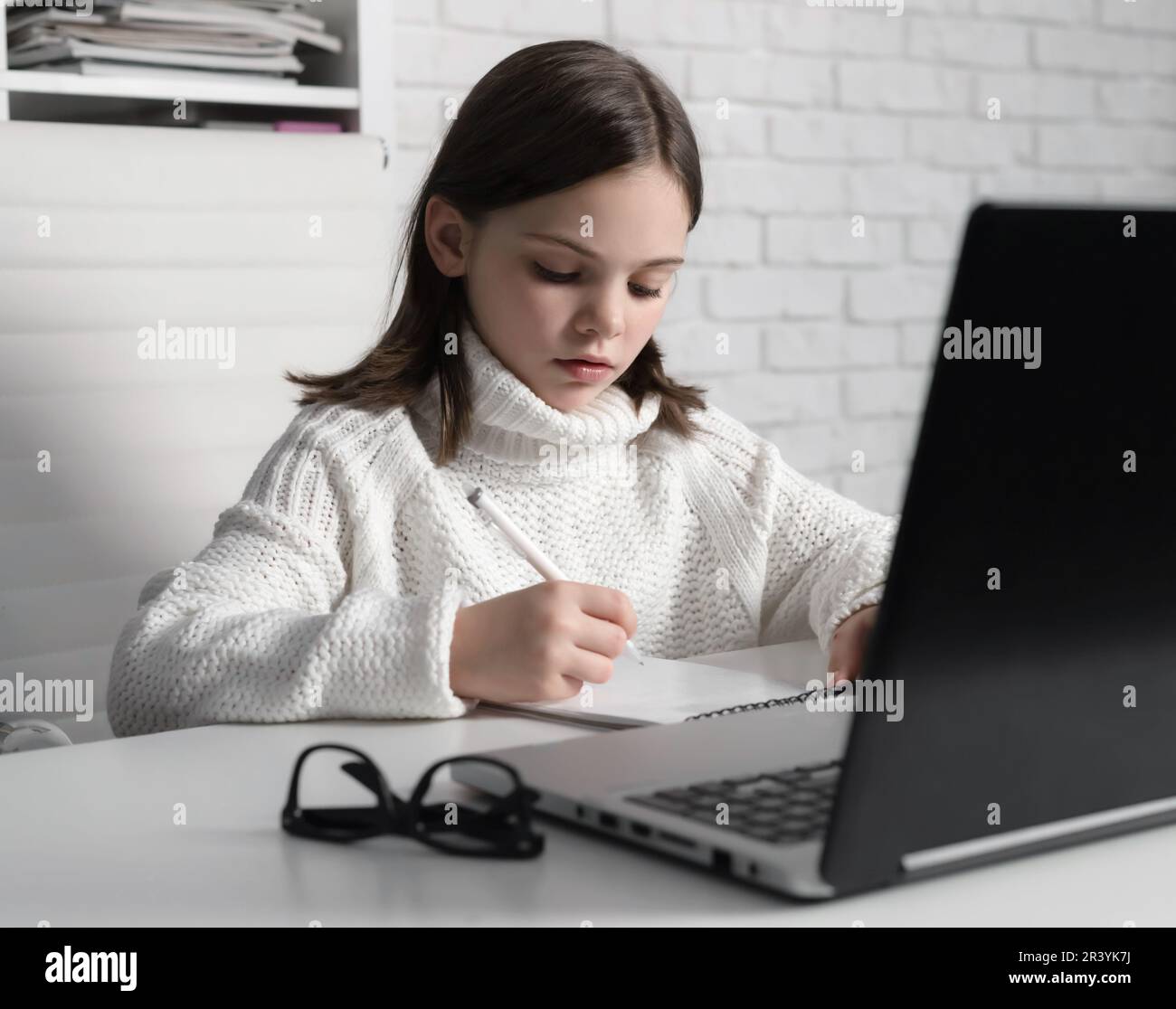 Child sitting at desk and writing in notebook near laptop. Serious and concentrated little girl studying. Eyeglasses on white table. Online education Stock Photo