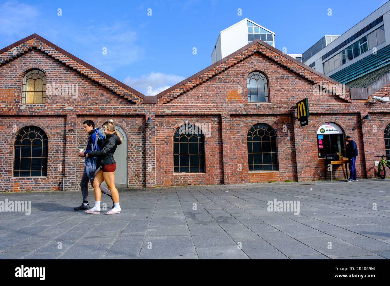 Sandnes, Rogaland, Norway, May18 2023, Young Couple Walking In Downtown Sandnes In Evening Sunlight Under A Blue Sky Passing Mcdonalds Hamburger Store Stock Photo