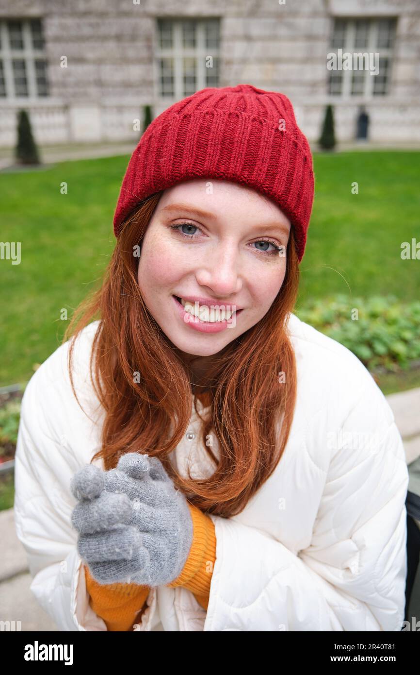 Vertical portrait of cute redhead female student in red hat and warm gloves,  sits in park on bench, smiles and looks cute at cam Stock Photo - Alamy