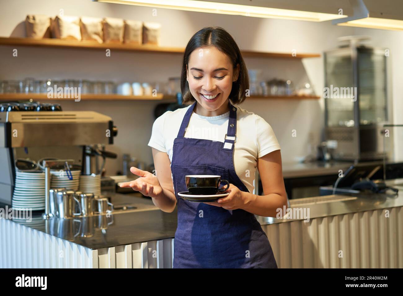 Portrait of smiling asian female barista, making coffee, holding cup of tea and taking it to cafe client, wearing apron, standin Stock Photo