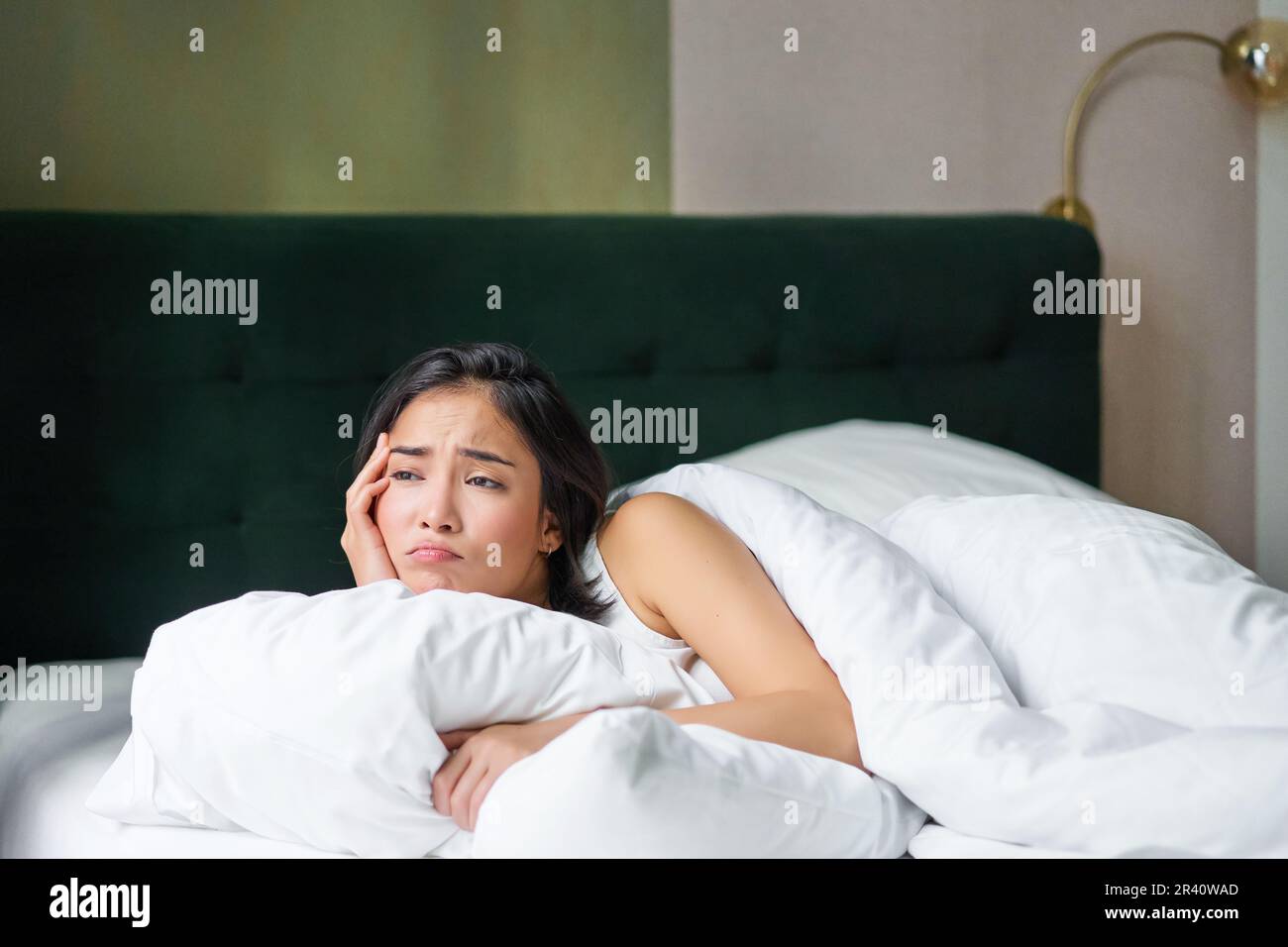 Portrait of upset korean woman, asian girl lying in bed feels sad, covers herself with duvet, looks outside window with disappoi Stock Photo