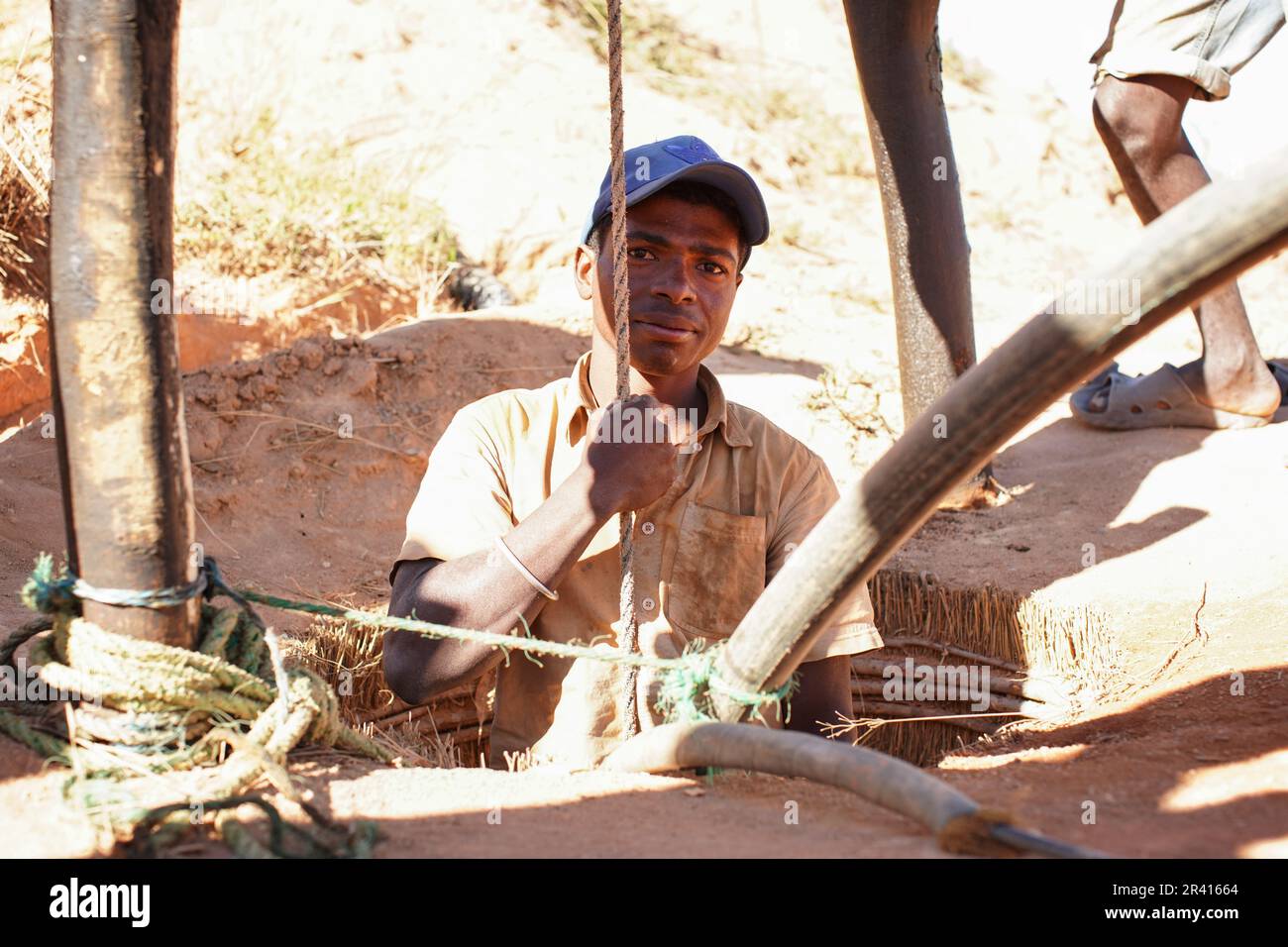 Ilakaka, Madagascar - April 30, 2019: Unknown Malagasy man standing in ground precious stone mine hole or well, half body visible, holding rope - abou Stock Photo