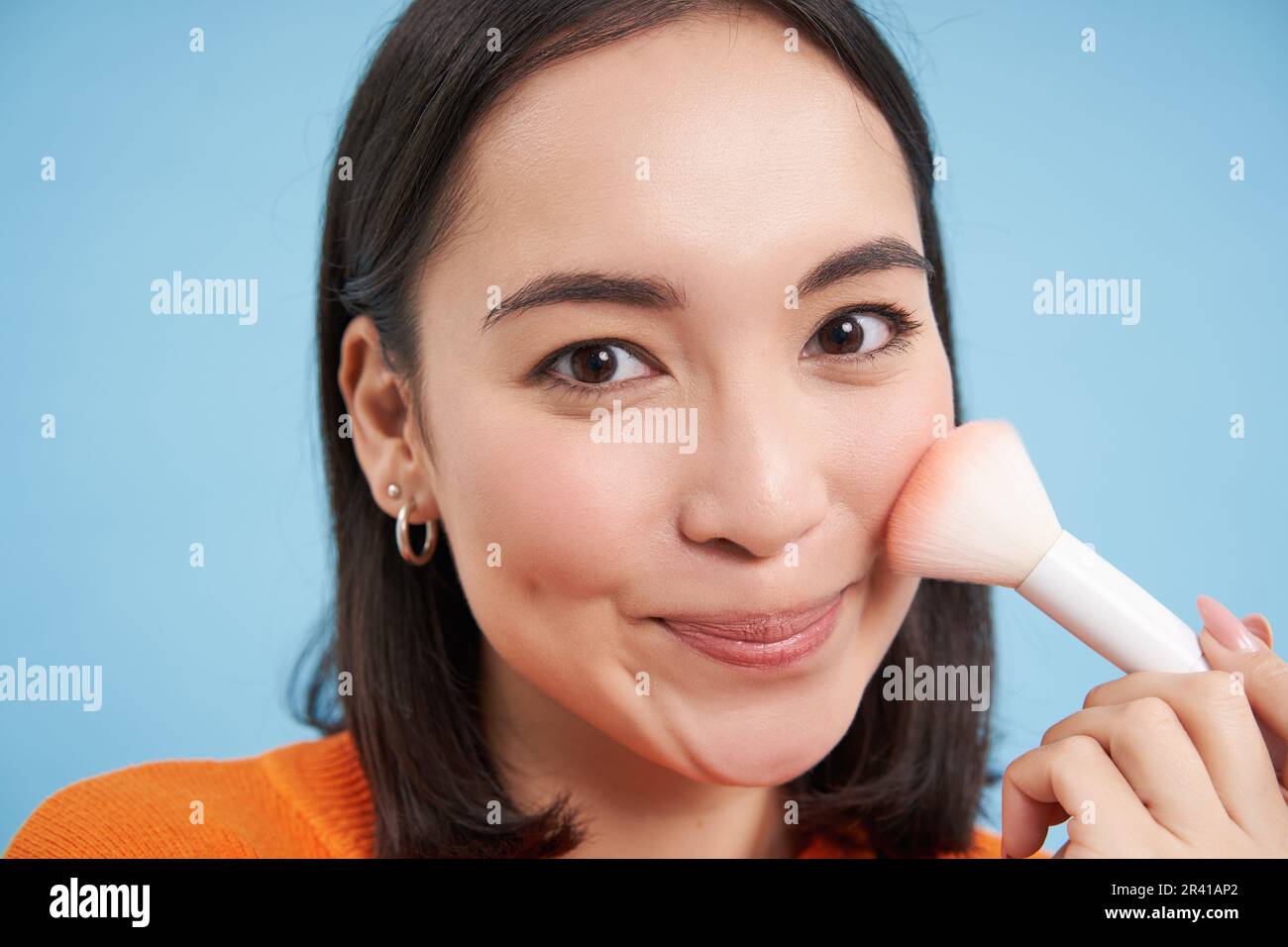 Makeup and beauty. Cute asian woman using cosmetic brush, puts on blush or  highlighter, stands over blue background Stock Photo - Alamy
