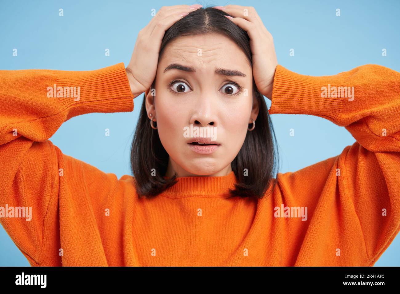 Close up of shocked asian woman in panic, anxious, looks terrified, stands over blue studio background Stock Photo