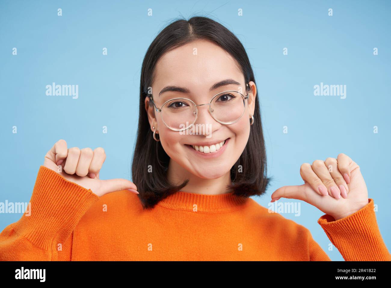 Portrait of cute korean woman in glasses, pointing fingers at camera self-assured, smiles with confidence, blue background Stock Photo
