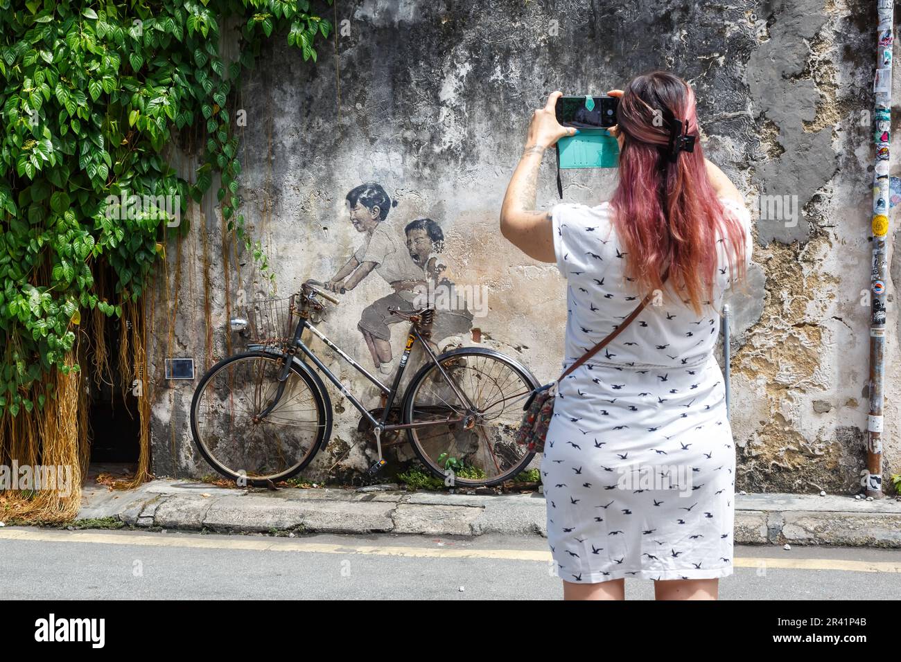 Penang, Malaysia - 7. Februar 2023: Junge Frau macht ein Foto von Street Art Kunst Junge und Mädchen auf Fahrrad an einer Mauer in George Town auf der Stock Photo