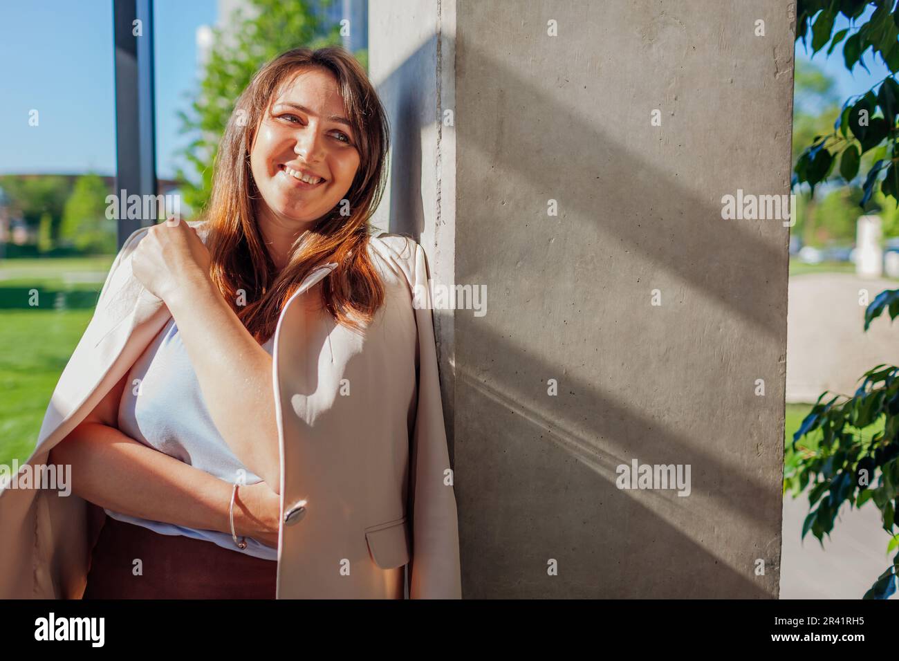 Happy businesswoman standing by wall in sunny office. Young smiling boss manager relaxing. Woman wears suit Stock Photo
