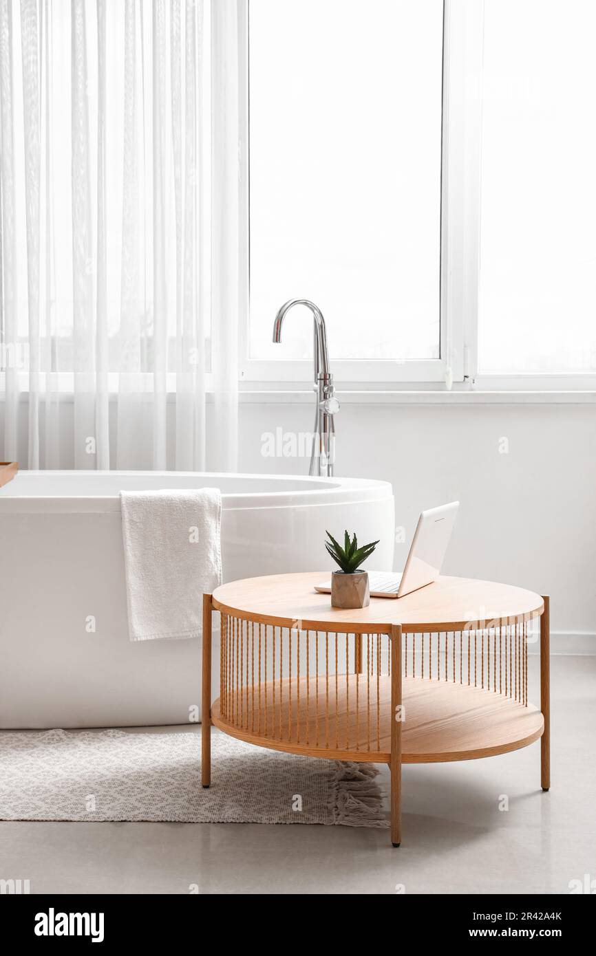 Modern laptop on coffee table and bathtub in interior of bathroom with